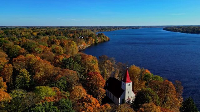 Aerial view of a church with a red roof surrounded by autumn foliage near the Daugava river in Kegums, Ogres novads, Latvia.