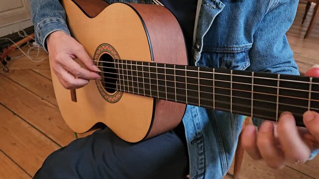 A close-up of a musician s hands playing an acoustic classical guitar at home, focusing on finger placement, the wooden floor, denim jacket, and the intimate atmosphere of practice and concentration.