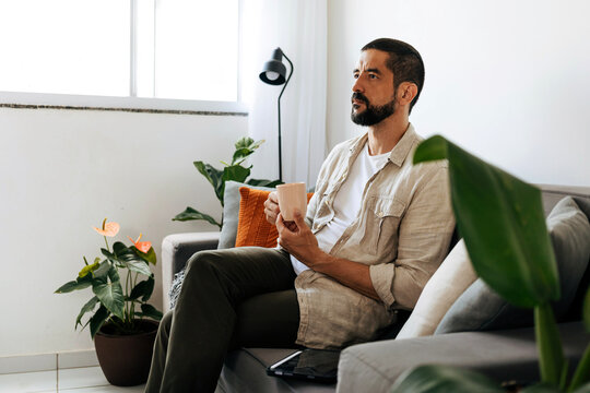 Thoughtful Man Sitting On Sofa With Mug In Modern Living Room Surrounded By Plants And Natural Light