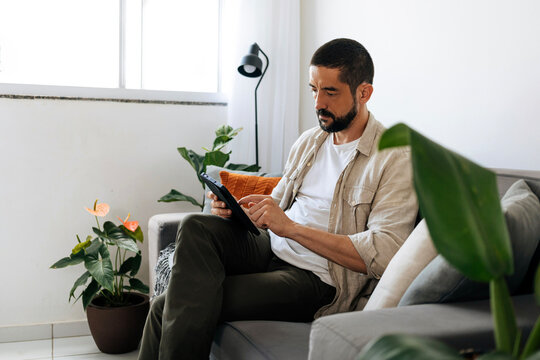 A focused man sits on a sofa at home using a tablet for work or
