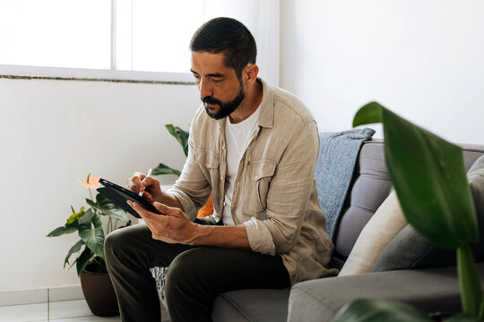 A focused man sits on a sofa at home using a tablet for work or leisure. Natural light, casual clothing and indoor plants create a relaxed, modern living room atmosphere.