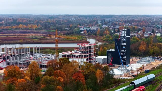 Aerial view of construction site with a tall crane, unfinished concrete structures, and industrial buildings near a river and railway tracks in Jelgava, Latvia.