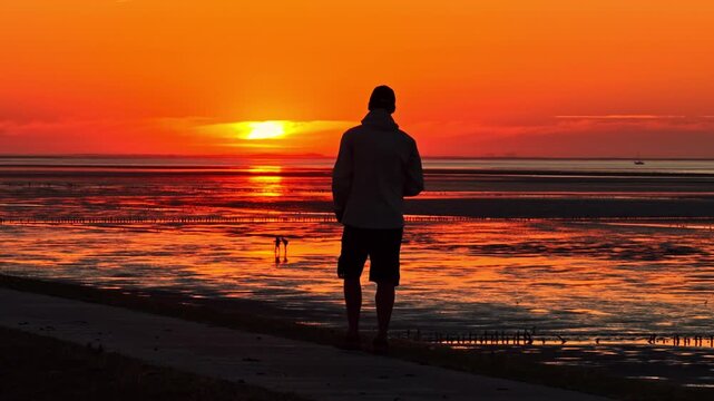 Silhouetted man photographing a radiant orange sunset over the tidal flats along the Netherlands coast - Aerial slow motion shot