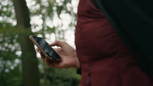 Handheld medium close up of a woman holding and using her smartphone while walking through a forest, with her face out of frame and green trees in the background.