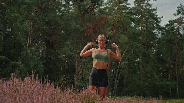 Fit athletic Caucasian woman wearing sportswear and headphones performs an upward arm and shoulder stretch in nature, then stretches her quadriceps by lifting and holding each foot behind her.