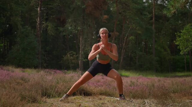 Slow motion footage of an athletic Caucasian woman in sportswear performing Cossack squats in nature with headphones, combining outdoor exercise and strength training among purple heather and forest.