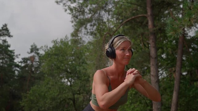 Medium shot of an athletic woman with defined abs wearing headphones as she performs Cossack squats in nature, focused and enjoying her outdoor workout.