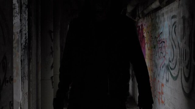Threatening figure carrying a wooden stick through dark abandoned factory graffiti-covered corridor, creating an atmospheric scene of fear, suspense and mystery