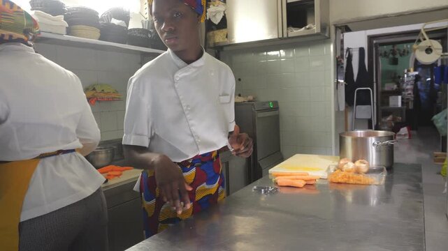 Female chef prepares ingredients beside vegetables and food containers on a stainless steel counter while another chef works nearby, medium shot