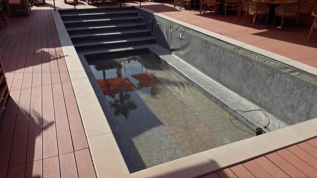 A high angle shot shows a rectangular swimming pool with stone steps surrounded by a wooden deck and outdoor dining furniture