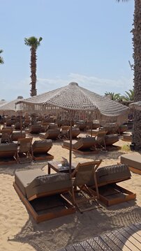 Panoramic view of a sandy beach club featuring woven fringed umbrellas wooden sun loungers and palm trees under bright daylight