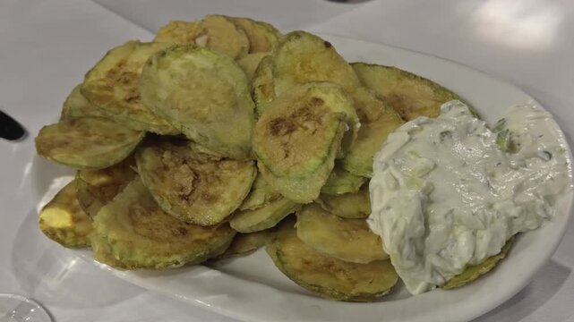 A white plate holds a stack of golden fried zucchini chips beside a scoop of tzatziki sauce ready to serve
