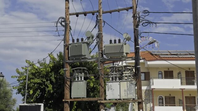 Electrical utility poles with transformers stand beside a multi story apartment building and green trees under blue sky