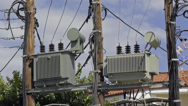 Two grey electrical transformer units are fitted on wooden power poles with wires crossing above red tiled rooftops