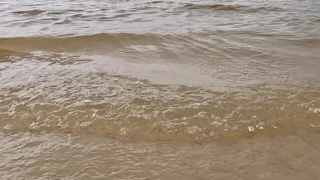 a wave crashes onto a sandy shore, the edges of the foam advance and recede rhythmically, and the water rises forward over the wet sand, creating light ripples along the shoreline in daylight.