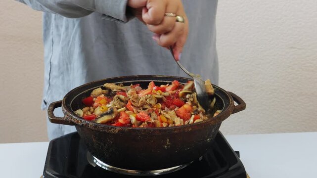 Vegetables and meat are stewed in a deep cast-iron skillet, with a female hand stirring with a spoon. Medium shot.