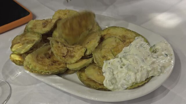 A plate of crispy fried zucchini slices is served with a side of creamy tzatziki dip and a fork at a Greek restaurant table
