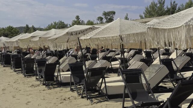Long rows of empty sun loungers under fringed umbrellas stretch along the sand with a forested backdrop at a beach club in Chalkidiki Greece