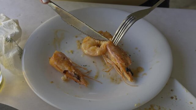 A hand uses a fork to eat a cooked shrimp on a white plate viewed from above during a seafood meal in Chalkidiki Greece