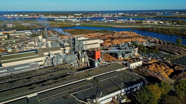 Aerial view of industrial facility with large factory buildings and wood chip piles located near the river in Riga, Latvia.
