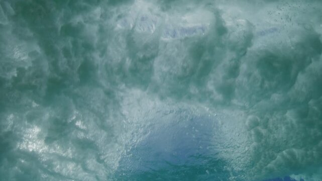 Sunlit breaking wave rolls overhead through clear turquoise ocean water with white foam below, clouds and sun hide behind whitewash