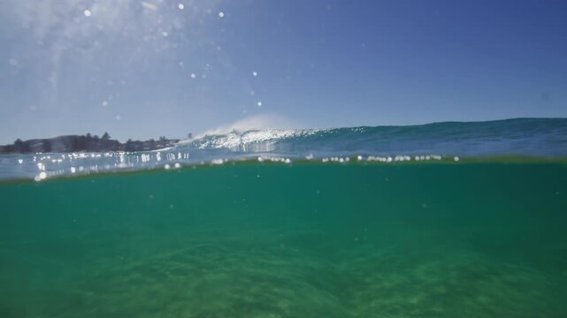 Ocean wave rolls through clear turquoise water beneath bright sunlight near a distant shoreline, splitlevel 50-50