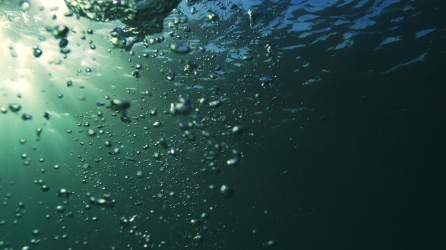 Air bubbles rise toward a rippled sunlit surface through clear blue green water underwater, space for text with subtle blue glow and stunning rays dancing