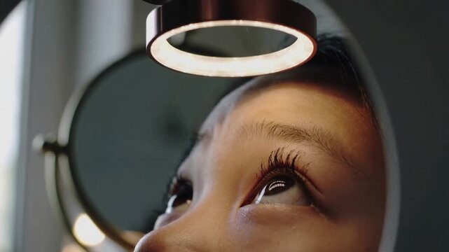 Lowering ring light drawing child gazing upward at round vanity mirror, casting warm corneal glint
