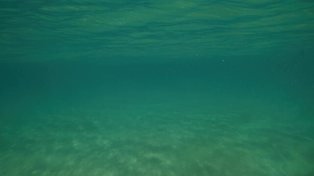 Clear turquoise ocean water reveals a sandy bottom beneath soft ripples moving across the surface