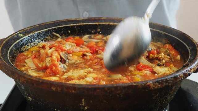 In a deep cast iron skillet, vegetables are stewed with chicken and mushrooms in sauce and spices, stirred with a spoon. Medium shot