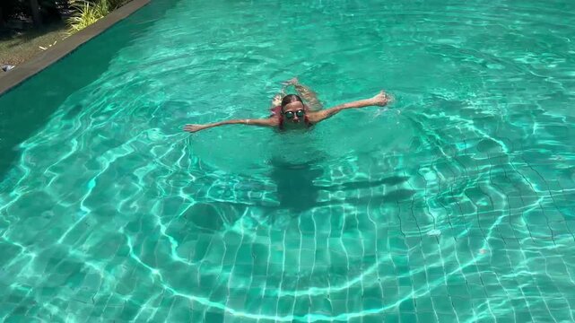 Woman swims in clear pool, making waves as they move. The sun shines brightly on water, reflecting light and creating a playful look. It is a warm day.