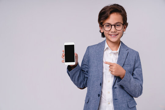 Cheerful caucasian boy pointing at blank smartphone screen isolated on grey background, childhood, technology, communication, digital lifestyle and presentation concept