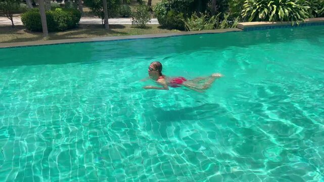 Woman swims in clear pool, making waves as they move. The sun shines brightly on water, reflecting light and creating a playful look. It is a warm day.