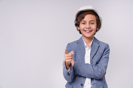 Cheerful small male kid in white hardhat pointing at camera isolated on grey background, childhood, construction, engineering, future profession, career and builder concept