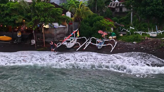 Aerial view of Amed Beach with traditional outrigger boats on the black sand shore and people walking near the water in Bali, Indonesia.