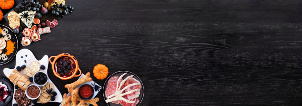 Halloween theme grazing corner border against a black wood banner background. Spooky charcuterie boards of meat and cheese appetizers. Above view with copy space.