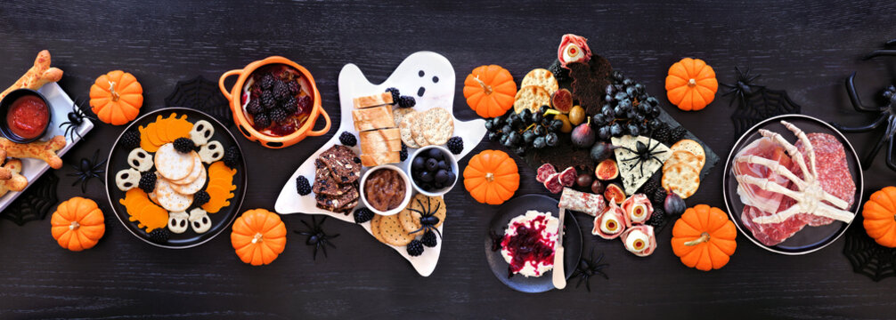 Halloween theme grazing table scene against a black wood banner background. Spooky charcuterie boards of meat and cheese appetizers. Overhead down.