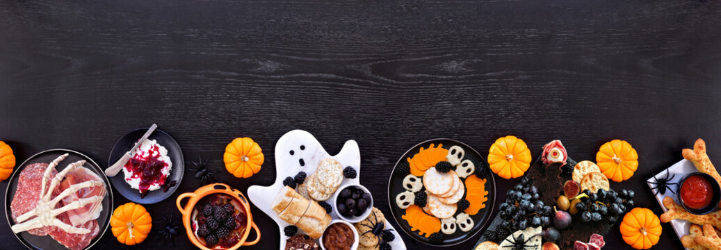 Halloween theme grazing bottom border against a black wood banner background. Spooky charcuterie boards of meat and cheese appetizers. Top view with copy space.