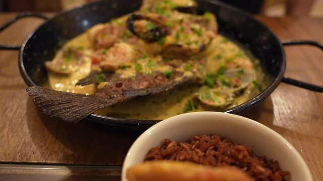 Close-up shot of a vibrant Colombian seafood stew in creamy sauce, garnished with herbs and served with red rice and patacones on rustic wooden table under warm indoor lighting.