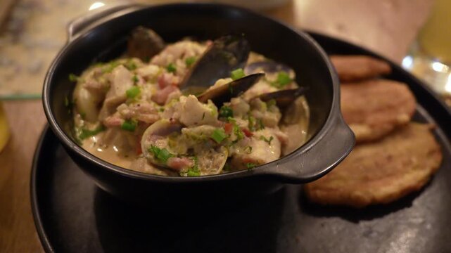 Appetizing close-up shot of creamy Colombian seafood stew garnished with fresh herbs, served in a black bowl beside crispy patacones on a dark plate under cozy, ambient lighting.