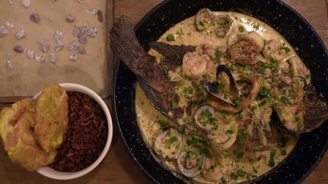 Overhead close-up shot of a richly garnished Colombian fish and seafood stew in creamy sauce, served with patacones and coconut rice on a rustic wooden table under warm lighting.