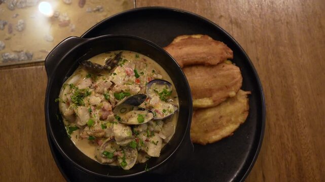 Overhead close-up shot of a traditional Colombian seafood stew served in a black bowl, garnished with fresh herbs and paired with golden patacones on rustic wooden table under warm lighting.