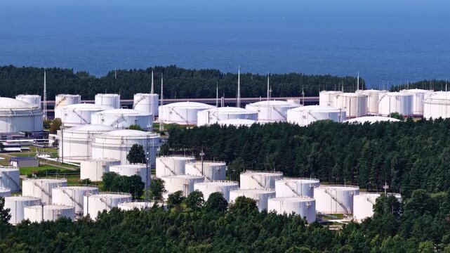 White storage tanks spread through industrial gas oil refiner holding complex beside forest and blue sea, aerial