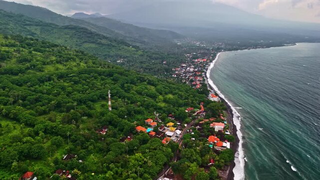 Aerial view of Amed Beach with lush green hills, coastal village, and black sand shoreline in Bali, Indonesia.