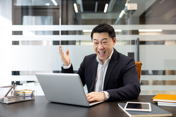Fototapeta premium Asian businessman working on his laptop, smiling and expressing excitement with a raised hand, celebrating victory, success, and achievement in his modern office workspace