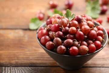 Fresh red gooseberries in bowl and green leaves on wooden table, closeup. Space for text © New Africa