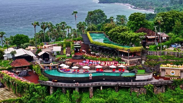 Aerial view of Amed Beach resort featuring a tiered infinity pool overlooking the ocean surrounded by lush tropical greenery and palm trees in Bali, Indonesia.