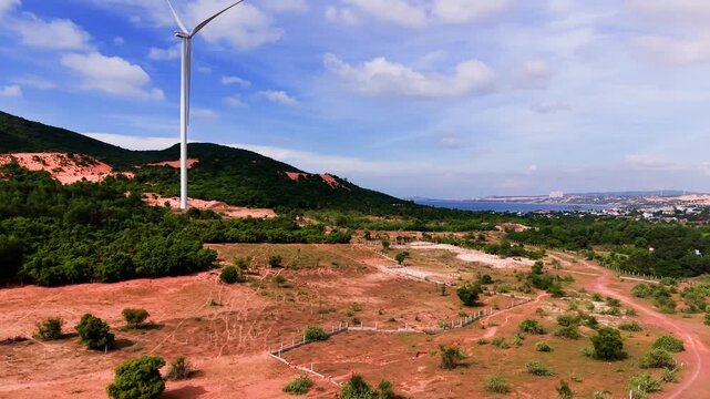 This shows a panoramic view of Hoa Thang in Lam Dong. Wind turbines stand tall against a backdrop of hills and open land. The skies are blue with scattered clouds above the scenery.