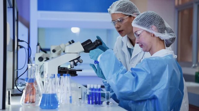Two female scientists in protective lab coat and hairnet examine a sample through a microscope, surrounded by flask and test tube filled with blue liquid in a laboratory research setting