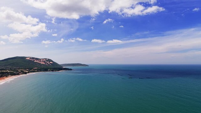 The camera shows wide shots of the landscape in Hoa Thang, Lam Dong. The clear sky meets the blue water of the coast. Small waves roll onto the shore. The area has hills in the background.